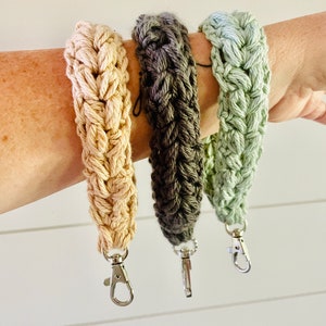 May include: Three braided wrist straps in beige, gray-brown, and green. Each strap is made of thick yarn and has a silver-colored lobster clasp. The straps are displayed against a white background.