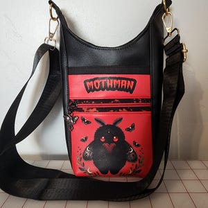 Może przedstawiać: Czarna torebka na ramię z czerwonym panelem przednim z napisem MOTHMAN. Torebka ozdobiona jest grafiką puszystego czarnego stworzenia z czerwonymi oczami trzymającego serce, otoczonego mniejszymi ćmami, listowiem i zawieszką suwaka w kształcie ćmy.