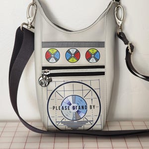 May include: A light gray crossbody bag with a black strap and silver hardware. The bag features a retro television test pattern design with the text "PLEASE STAND BY." It has a zipper pocket and three colorful circular graphics.