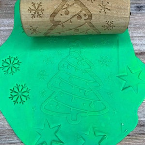 May include: A wooden rolling pin with a Christmas tree and snowflake design is used to create impressions in green play dough. The dough has been rolled out and shows the impressions of the rolling pin, including a Christmas tree, snowflakes, and stars.