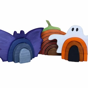 May include: Three wooden Halloween-themed rainbow stackers. The first is a purple bat with gray and blue arches. The second is an orange and brown pumpkin with green stem. The third is a white ghost with orange and brown arches.