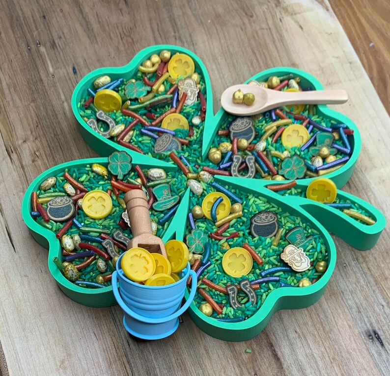May include: A green shamrock-shaped tray filled with green, yellow, and gold sprinkles, gold coins, and other St. Patrick's Day themed decorations. A small wooden scoop with a blue bucket filled with gold coins sits in the tray.