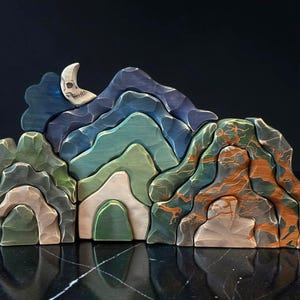 May include: A multi-piece wooden stacking toy shaped like a mountain range with textured surfaces. The pieces feature shades of green, blue, brown, and cream, with a crescent moon shaped piece at the top that has a carved skull design on its surface.