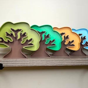 May include: Four colorful tree-shaped cookie cutters, made of plastic, in shades of green, blue, orange, and brown. The cutters are arranged in a row on a wooden shelf.