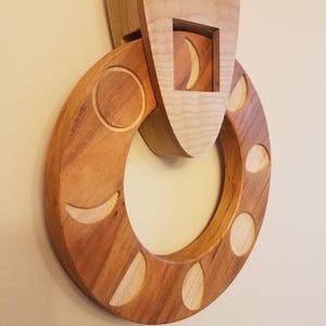 May include: A wooden wall hanging with a circular design featuring various phases of the moon carved into the wood. The moon phases are arranged in a circle around a central opening. The piece is made of light brown wood and has a rustic, handcrafted look.