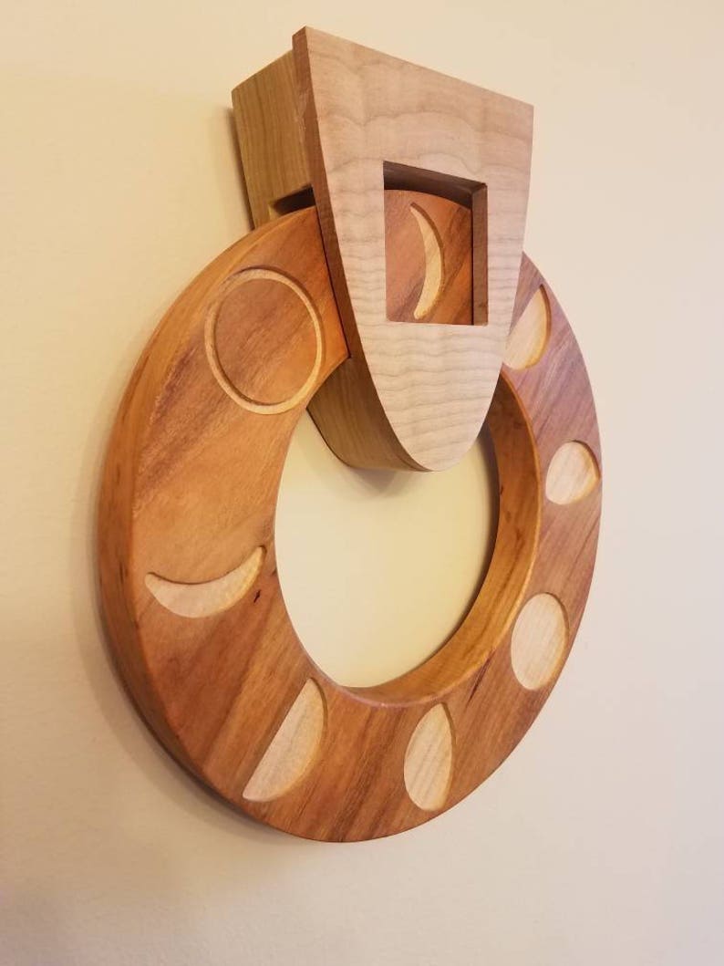 May include: A wooden wall hanging with a circular design featuring various phases of the moon carved into the wood. The moon phases are arranged in a circle around a central opening. The piece is made of light brown wood and has a rustic, handcrafted look.