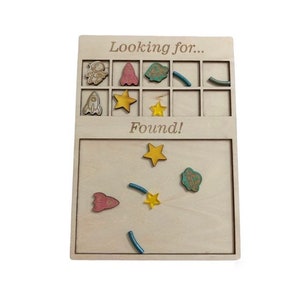 May include: Wooden game board with a grid of squares. The top section says "Looking for..." and the bottom section says "Found!" There are small wooden pieces with images of rockets, stars, and astronauts. Some pieces are in the grid and some are on the bottom section.