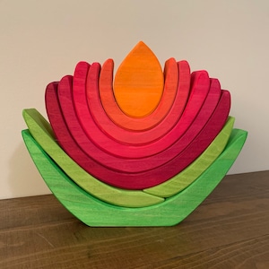 Puede incluir: Una flor de juguete de madera con un esquema de colores rojo, naranja y verde. La flor está compuesta por arcos apilados y una sola pieza en forma de lágrima.