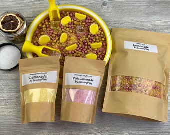 Può includere: Una ciotola gialla piena di riso sensoriale giallo e rosa, due palette e un barattolo di frutta di agrumi secca. Tre sacchetti di carta marrone con finestre trasparenti contengono polvere sensoriale gialla e rosa. I sacchetti sono etichettati "Sensory Fizzy Powder Lemonade By Sensory Play" e "Sensory Fizzy Powder Pink Lemonade By Sensory Play".