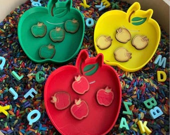 Può includere: Tre vassoi a forma di mela in verde, giallo e rosso, ciascuno con ritagli di mele in legno. I vassoi sono circondati da riso colorato e lettere dell'alfabeto, creando un tema educativo giocoso.