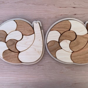 May include: Two wooden snail puzzles with a light brown and white color scheme. Each puzzle is made up of multiple pieces that fit together to form the shape of a snail.