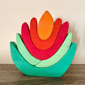 Puede incluir: Un juguete de apilar de madera con forma de flor de cinco pétalos. Los pétalos son de color verde, rojo y naranja. El juguete está hecho de madera natural y está pintado con pintura no tóxica.