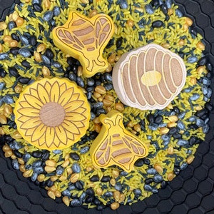 May include: A black bowl with a honeycomb pattern filled with yellow rice, black beans, and gold sprinkles. There are four wooden toys in the bowl: a bee, a honeycomb, a sunflower, and another bee.