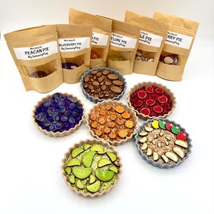 May include: A set of eight mini pie sensory play kits, each with a different flavor: pecan, blueberry, lime, apple, cherry. The kits come in brown paper bags with clear windows showing the contents. The pies are arranged in a circle on a white surface.
