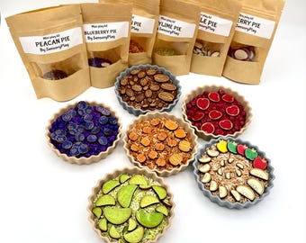 Può includere: Un set di otto mini kit di gioco sensoriale a base di torta, ognuno con un sapore diverso: pecan, mirtillo, lime, mela, ciliegia. I kit sono forniti in sacchetti di carta marrone con finestre trasparenti che mostrano il contenuto. Le torte sono disposte in cerchio su una superficie bianca.