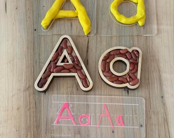 May include: A set of clear plastic trays with the letter A and a circle cut out. The letter A is filled with red kidney beans and the circle is filled with yellow playdough. There is a clear plastic sheet with the letter A written in pink marker.