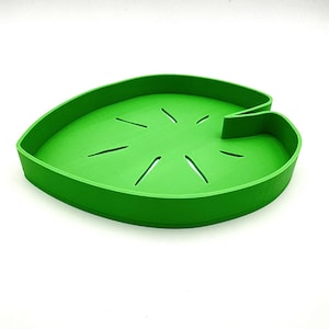 May include: A bright green, leaf-shaped tray with a raised edge and several linear cutouts. The tray is made of a solid material and has a smooth surface. The design mimics a lily pad, suitable for decorative or functional use.