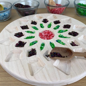 May include: A wooden tray with a circular design featuring multiple compartments. The compartments are filled with different colored rice, including green, red, and purple. A small wooden scoop is also included.