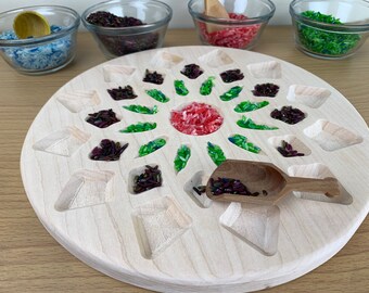 Op de afbeelding: Een houten dienblad met een cirkelvormig ontwerp met meerdere vakken. De vakken zijn gevuld met verschillende gekleurde rijst, waaronder groen, rood en paars. Een kleine houten schep is ook inbegrepen.