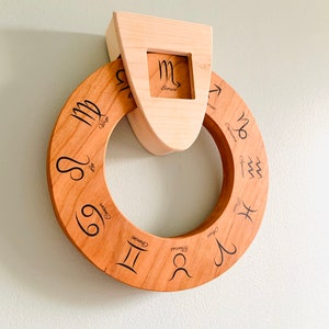 May include: A wooden zodiac wheel with the astrological signs and names engraved on it. The wheel is attached to a wooden frame with the Scorpio sign in a cutout.