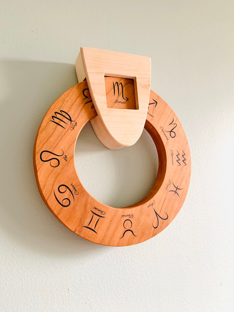 May include: A wooden zodiac wheel with the astrological signs and names engraved on it. The wheel is attached to a wooden frame with the Scorpio sign in a cutout.