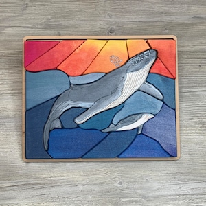 May include: A wooden puzzle featuring a whale and a smaller whale swimming in a blue and orange ocean. The puzzle pieces are shaped like the ocean and the whales.
