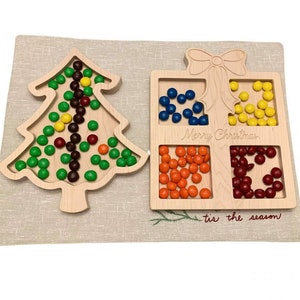 May include: Wooden Christmas tree and gift box shaped trays with colorful candies inside. The tree tray has green, brown, yellow, and red candies. The gift box tray has blue, yellow, orange, and red candies. The gift box tray has the text "Merry Christmas" and "'tis the season" engraved on it.