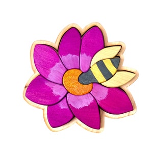 May include: A wooden puzzle of a pink flower with a bee landing on it. The flower is made of multiple pieces that fit together. The bee is also made of multiple pieces.