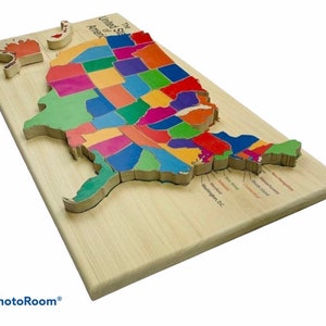 May include: A wooden puzzle of the United States of America, with each state a different color. The puzzle is on a light-colored wooden base, with some state pieces removed. The text "The United States of America" is visible.