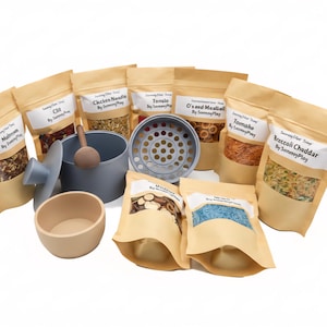 May include: A collection of toy food items, including pouches of various ingredients, a gray pot with a lid, a wooden spoon, and a small bowl. The pouches have labels with food names. The items are arranged on a white surface.