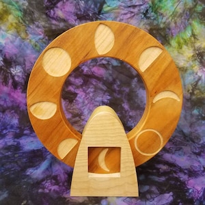 May include: Wooden moon phase wheel with a crescent moon in the center. The wheel has eight different moon phases carved into it. The wheel is made of light brown wood and is set on a wooden stand.