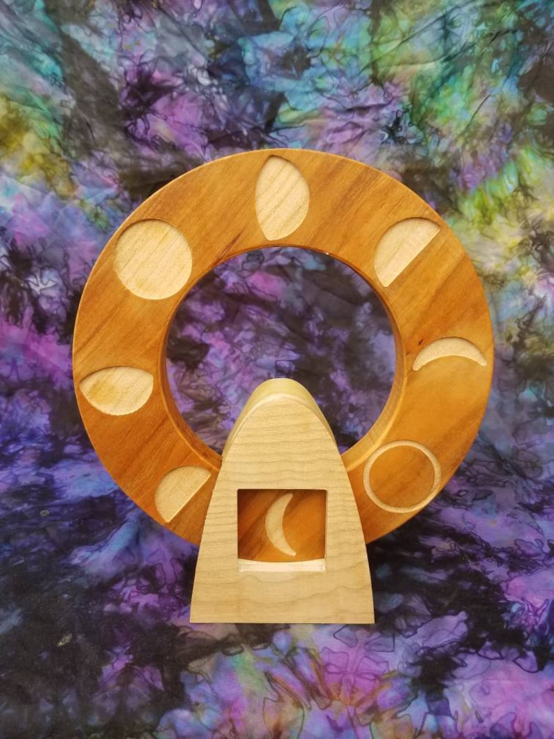 May include: Wooden moon phase wheel with a crescent moon in the center. The wheel has eight different moon phases carved into it. The wheel is made of light brown wood and is set on a wooden stand.