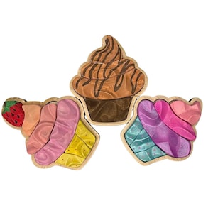 May include: Three wooden cupcake puzzles with colorful frosting and wooden bases. One has a strawberry on top, another has chocolate frosting, and the third has pink and blue frosting. The puzzles are designed for children.