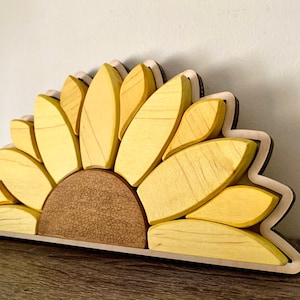 May include: A wooden sunflower wall decor with yellow petals and a brown center. The sunflower is made of individual pieces of wood that are assembled together.