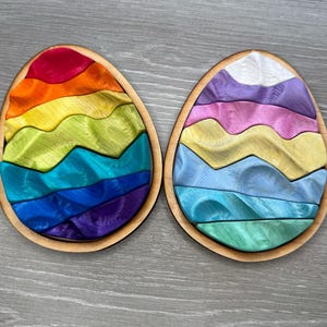 May include: Two wooden egg-shaped puzzles with a wavy design. One puzzle is rainbow colored and the other is pastel blue, green, and purple.
