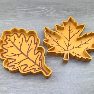 May include: Two decorative leaf-shaped trays, one shaped like an oak leaf and the other like a maple leaf. Both trays are a solid yellow color with brown outlines.