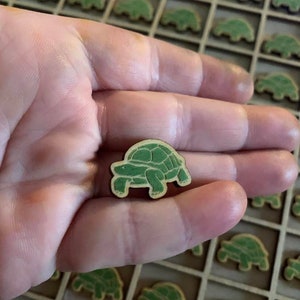 May include: A close-up of a small, green wooden turtle figurine. The turtle is being held in a person's hand. The turtle is surrounded by many other turtles, all of which are arranged in a grid pattern.