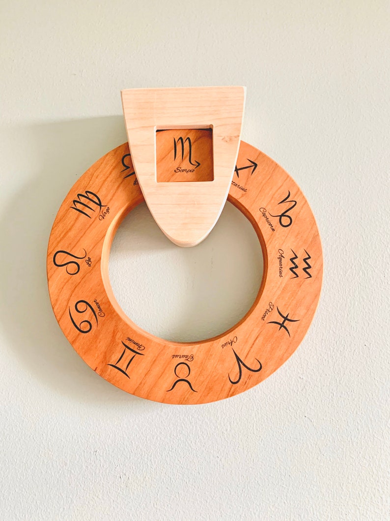 May include: A wooden zodiac wheel with the astrological signs engraved on it. The wheel is mounted on a wall with a separate wooden piece that displays the Scorpio sign.