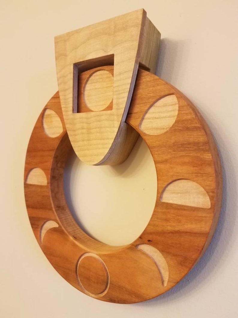 May include: A wooden wall hanging with a circular design featuring various phases of the moon. The moon phases are carved into the wood and are surrounded by a larger circle of wood. The top of the piece features a square cutout with a circle inside.