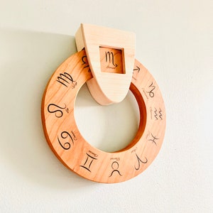 May include: A wooden zodiac wheel with the twelve astrological signs engraved on it. The wheel is mounted on a wooden plaque with a cutout for the sign Scorpio.