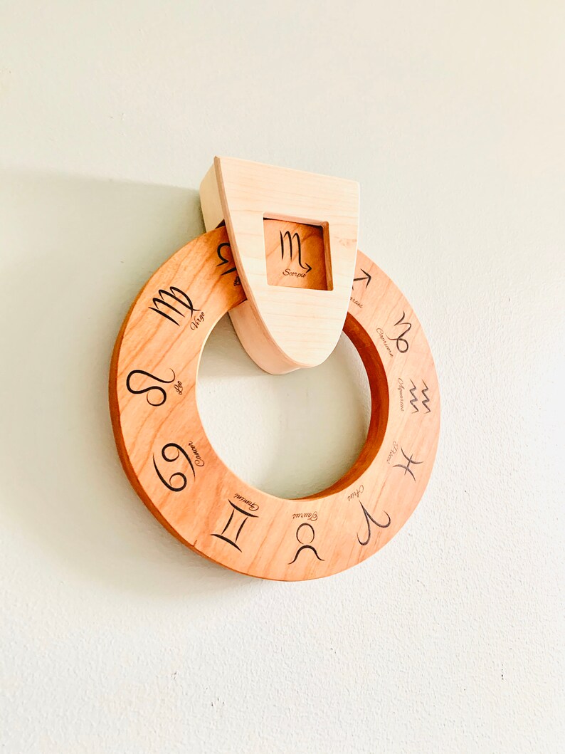 May include: A wooden zodiac wheel with the twelve astrological signs engraved on it. The wheel is mounted on a wooden plaque with a cutout for the sign Scorpio.