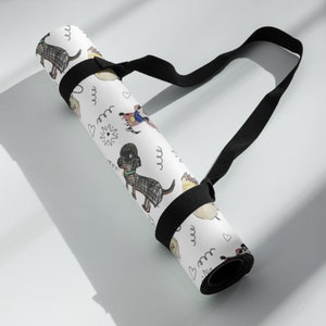 K&ouml;nnte beinhalten: Wei&szlig;e Yogamatte mit schwarzem Gurt. Die Matte hat ein sich wiederholendes Muster aus handgezeichneten Hunden und Herzen. Der Gurt ist mit einer schwarzen Schnalle an der Matte befestigt.