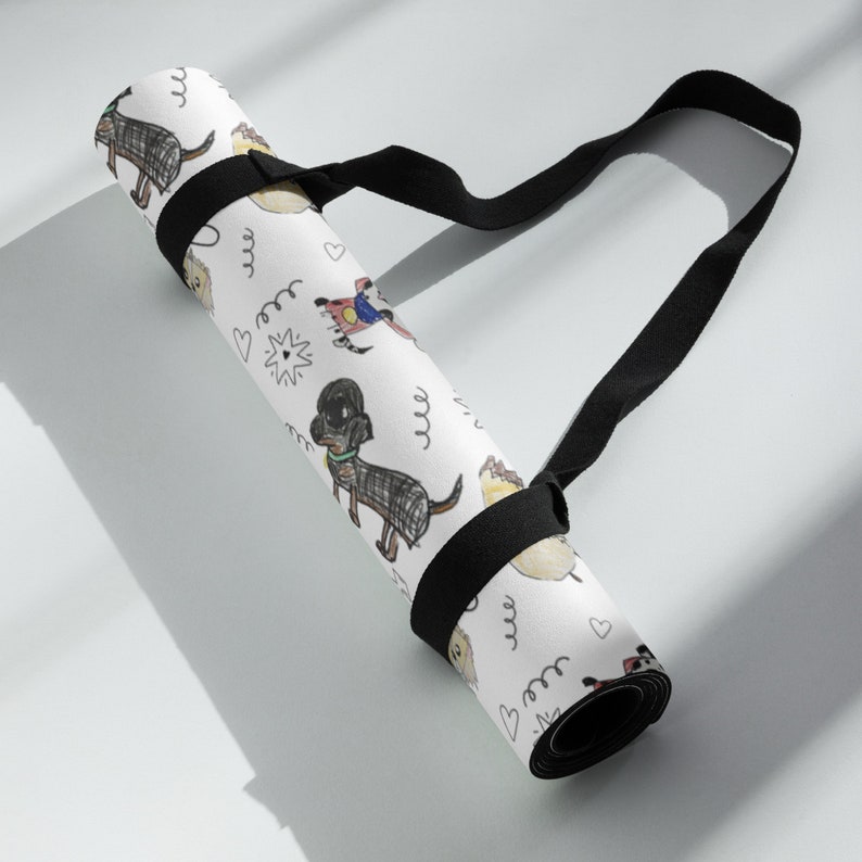K&ouml;nnte beinhalten: Wei&szlig;e Yogamatte mit schwarzem Gurt. Die Matte hat ein sich wiederholendes Muster aus handgezeichneten Hunden und Herzen. Der Gurt ist mit einer schwarzen Schnalle an der Matte befestigt.