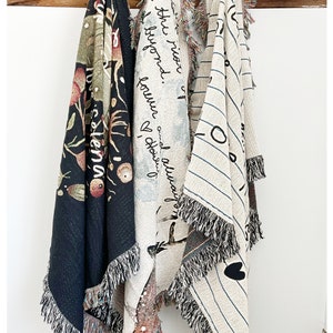 May include: Three woven tapestry blankets hanging on a wooden shelf. The blankets feature different designs, including a black and white striped pattern, a floral pattern, and a design with the text "and always found a reason to be beyond the moon".