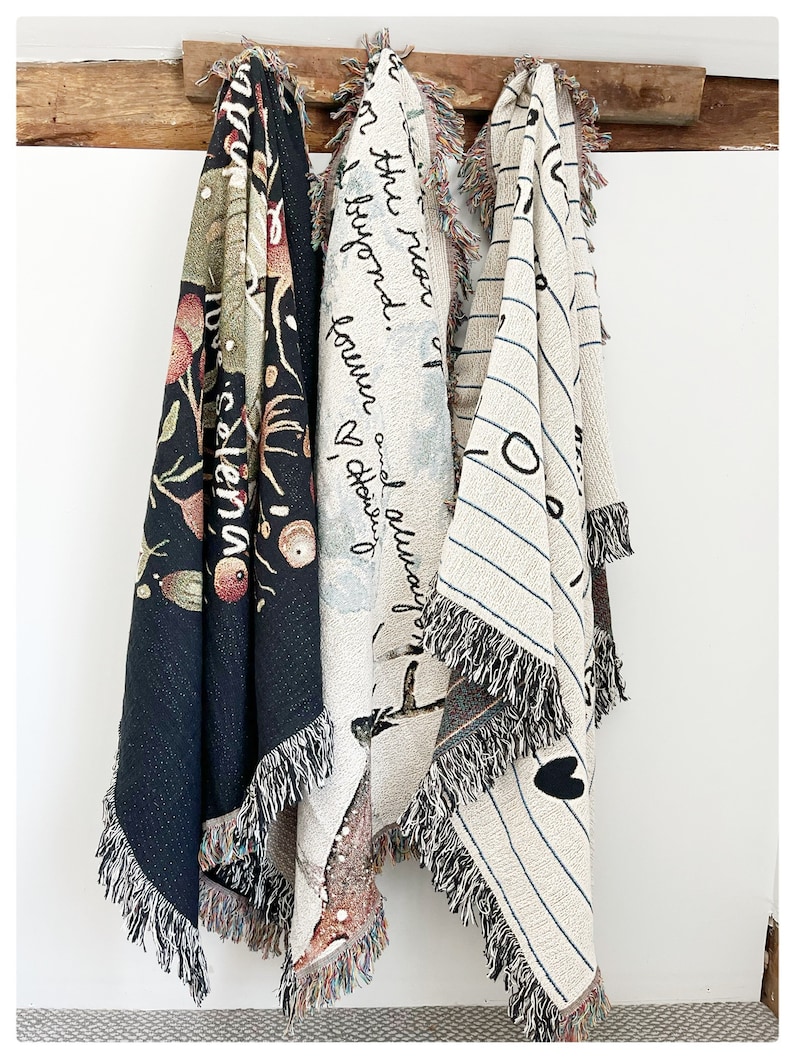 May include: Three woven tapestry blankets hanging on a wooden shelf. The blankets feature different designs, including a black and white striped pattern, a floral pattern, and a design with the text "and always found a reason to be beyond the moon".