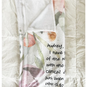 May include: A white throw blanket with a floral pattern in shades of pink, orange, and green. The blanket has a handwritten message that reads: "Audrey, I have of the N with the clinical has been who is so In that w"