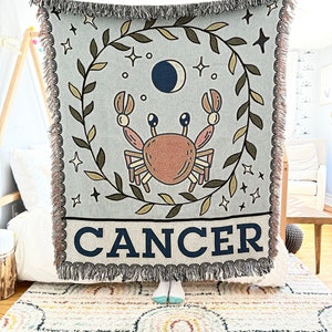 May include: A woven tapestry throw blanket with a blue background featuring a brown crab, a crescent moon, and stars. The blanket has a green leafy border and the word "CANCER" in black letters.