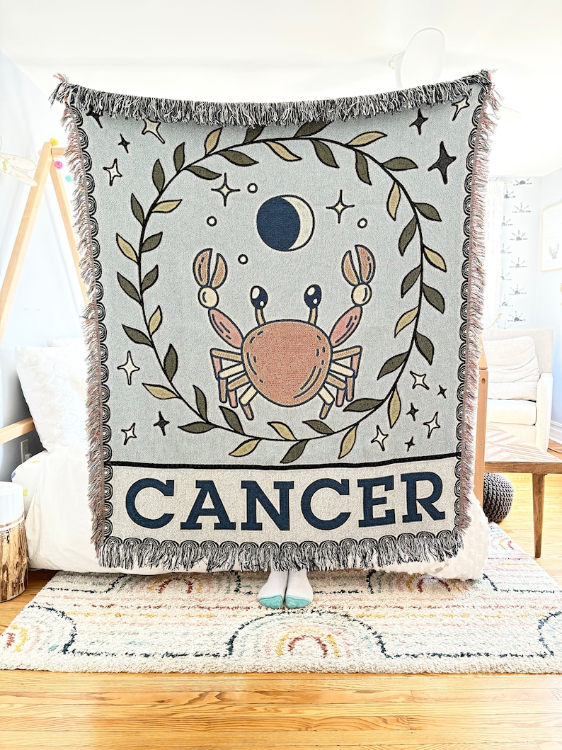 May include: A woven tapestry throw blanket with a blue background featuring a brown crab, a crescent moon, and stars. The blanket has a green leafy border and the word "CANCER" in black letters.