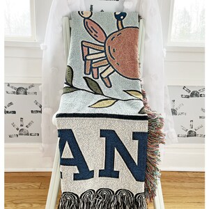 May include: A woven throw blanket with a blue background featuring a cartoon crab and the word "AN" in large, navy blue letters. The blanket has a fringe border.
