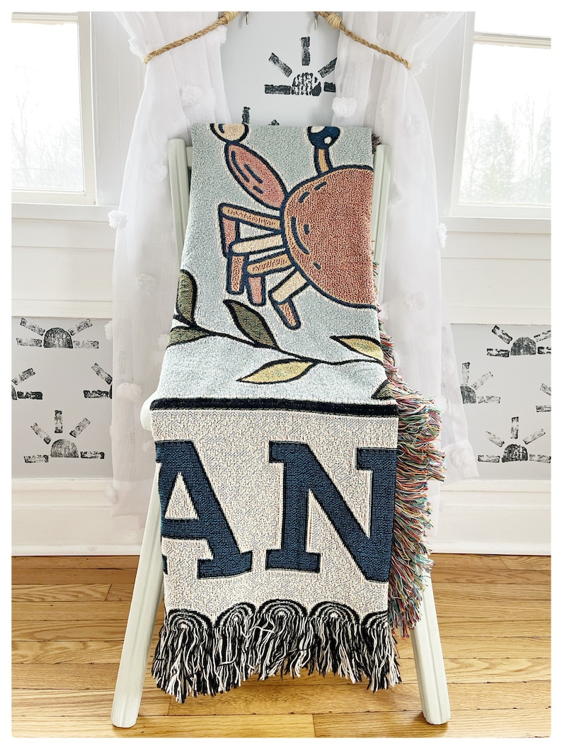 May include: A woven throw blanket with a blue background featuring a cartoon crab and the word "AN" in large, navy blue letters. The blanket has a fringe border.